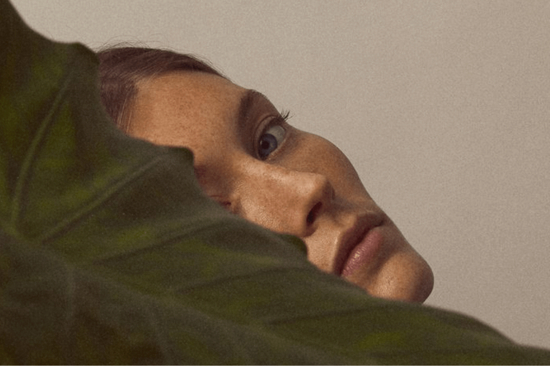 Close-up profile of a woman’s face partially obscured by a large green leaf, soft natural light highlighting skin texture and freckles.