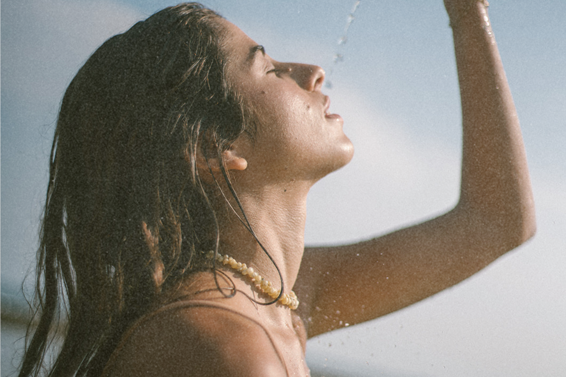Side profile of a person with wet hair outdoors, lifting their arm as water falls onto their face