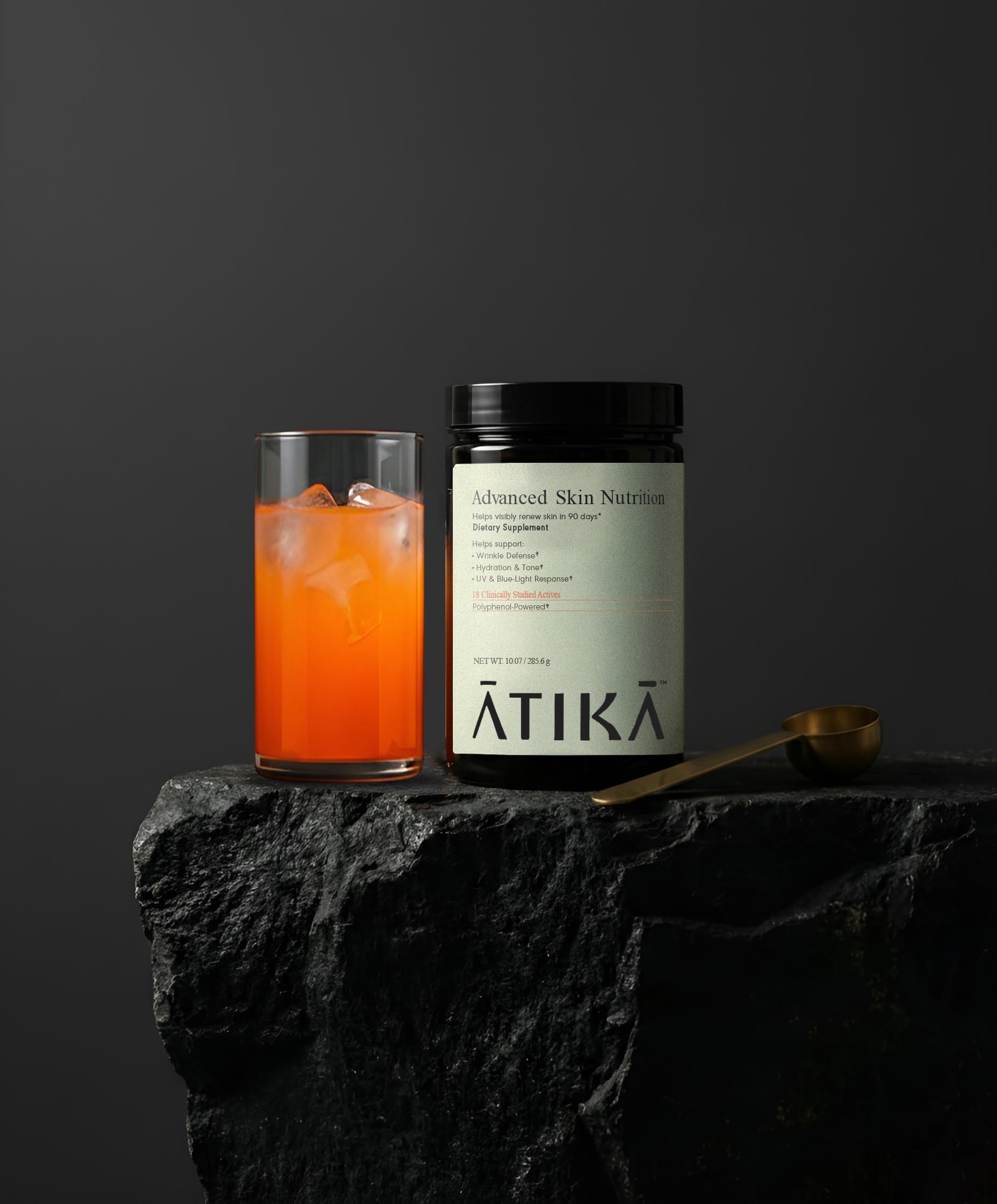 Jar of ATIKA Advanced Skin Nutrition on a dark surface with an orange beverage in the foreground.