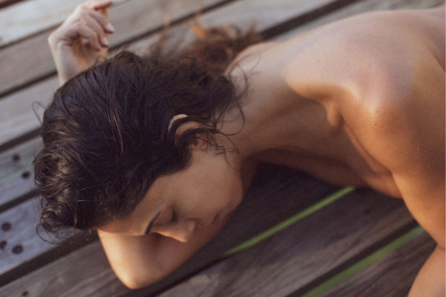 Woman lying on a wooden deck with bare shoulders, resting her head on her arm, showing natural sunlit skin.