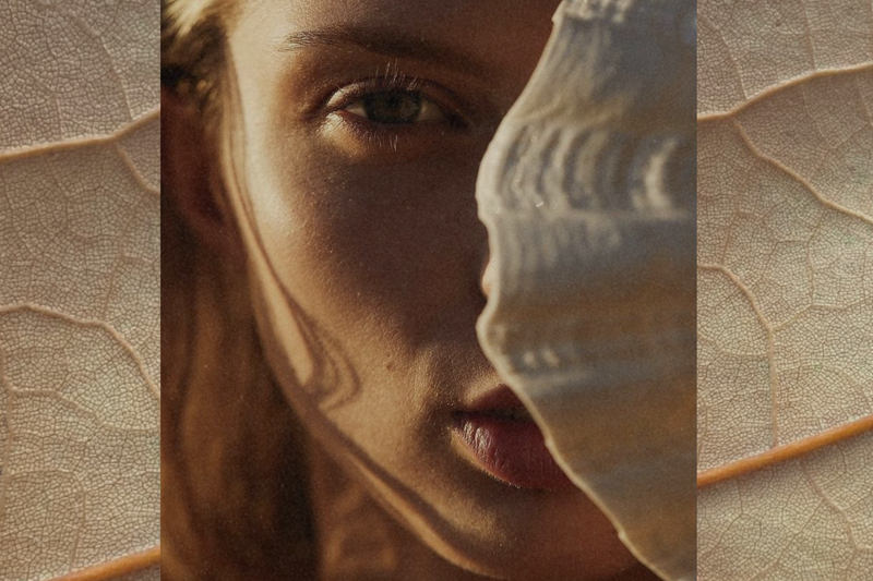 Sunlit close-up of a woman’s face partially veiled by a translucent leaf, warm light and organic textures creating a layered, natural feel.
