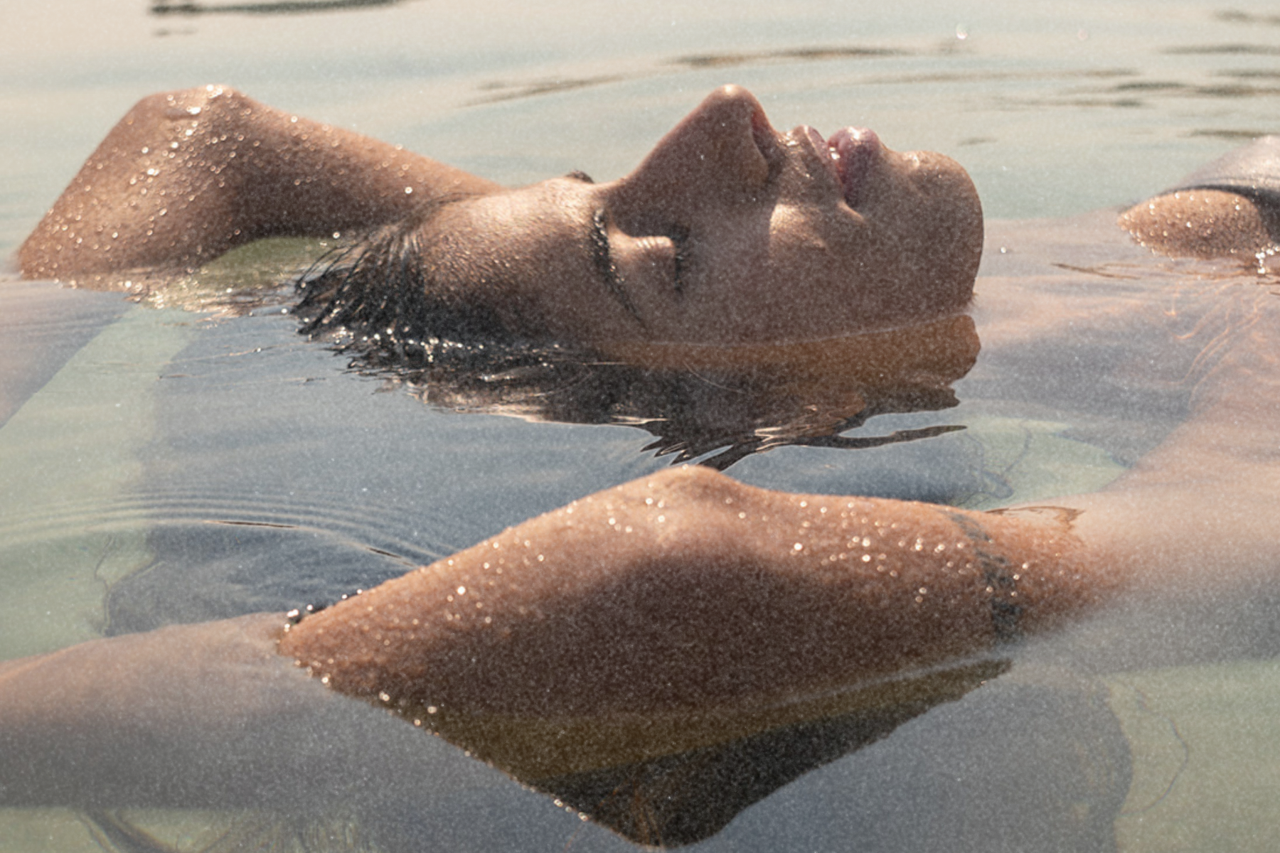 Person floating on their back in calm, shallow water with sunlight on the skin.